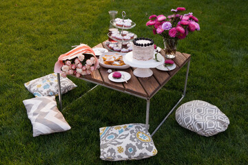Beautifully served table with desserts on green grass. Summer picnic in the garden with muffins, cookies, cake, fresh fruits, cold lemonade, wine and beautiful flowers with pink decorations.