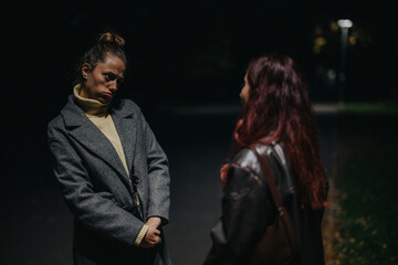 Two women interact in a nighttime outdoor setting, dressed warmly and expressing unique emotions. The scene conveys togetherness, casual communication, and a cozy ambiance.