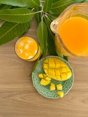 A fresh mango feast: a green plate holds diced and sliced ripe mango, next to a glass and pitcher of vibrant orange mango juice. Green mango leaves add a natural touch to the wooden table setting.