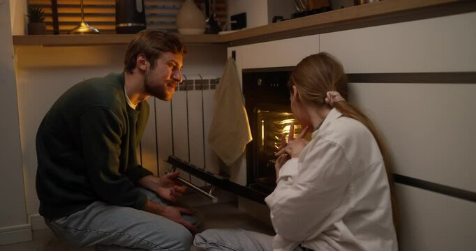 A happy guy and his girlfriend open the oven and inspect the finished dish tasting its smell while preparing dinner at home in the evening in the kitchen