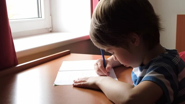 The child writes down the answer to the solution of the example in a copybook. A mathematics task. A boy diligently learns to write. The kid is sitting at the table and doing a homework. Smart person.