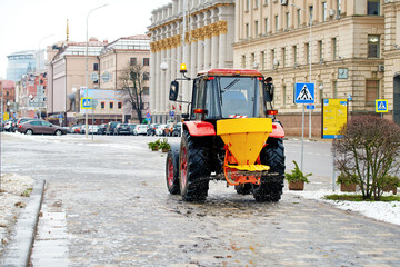 Tractor with salt spreader clearing icy road in city during winter, preventing accidents on slippery streets. Winter road maintenance in action, tractor spreads salt on frozen sidewalk. Spreading salt