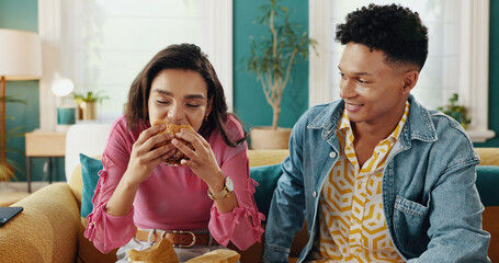 Eating, couple and delicious food for woman, home and people with meal in living room, smile or takeaway. House, happy and man in relationship with hungry partner enjoying taste of burger in lounge