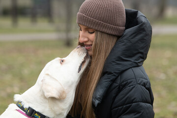 A woman wearing a knitted hat and puffy coat smiles as her white dog affectionately licks her face...