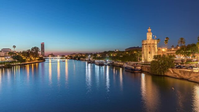 Tour boat floating along the Guadalquivir River near Torre del Oro watchtower in Seville, Spain. Day to night transition timelapse with city lights reflecting on water and scenic view of a waterfront