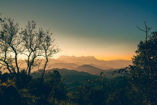 High-angle view of a beautiful warm sky in the evening as the sun sets. There is a view of Doi Luang Chiang Dao Mountain in the center, which has a unique shape. There are many trees in foreground.