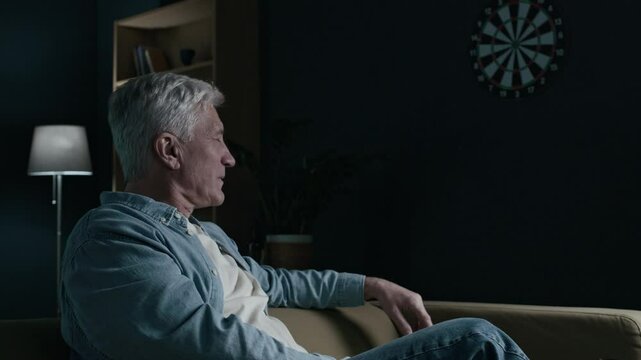 Low key shot of mature Caucasian man sitting on couch at home in front of beer bottles, throwing darts at a dartboard on wall in despair, then drinking another bottle of bear looking off into space