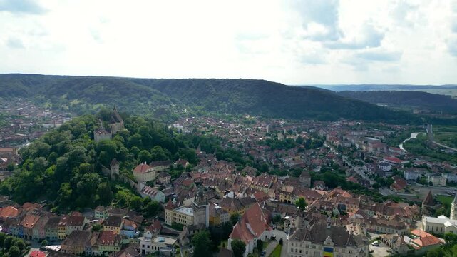 Aerial view of Sighisoara historic city center. Popular tourist destination and UNESCO World Heritage Site. Green landscape. City surrounded by forest. Drone going forward above the city center.