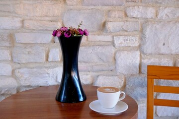 A black vase with pink flowers on the table. Coffee latte and wooden chair. Cafe culture. White tumbled stone wall in the background.