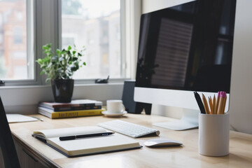 desk setup with monitor, notebook, pen, and potted plant near a window
