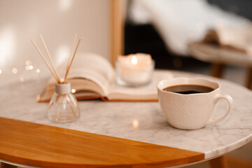 Cup of coffee with aroma diffuser and burning candle on paper book staying on marble table in room close up. Cozy home atmosphere.
