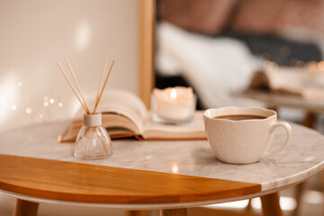 Cup of coffee with aroma diffuser and burning candle on paper book staying on marble table in room close up. Cozy home atmosphere.