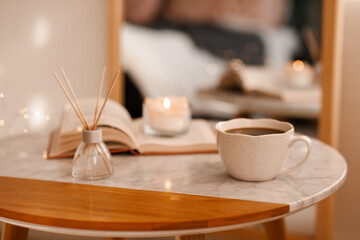 Cup of coffee with aroma diffuser and burning candle on paper book staying on marble table in room close up. Cozy home atmosphere.