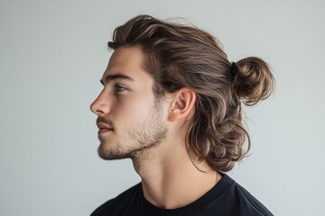 Close-up profile of a young man with long brown hair tied in a bun, highlighting his facial features and hairstyle against a simple background