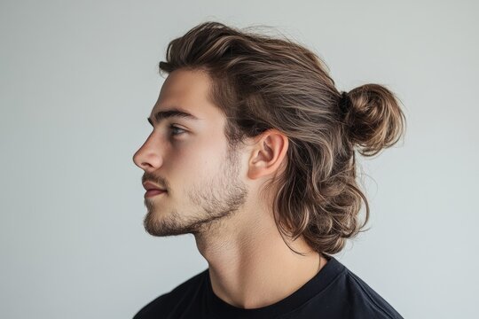 Close-up profile of a young man with long brown hair tied in a bun, highlighting his facial features and hairstyle against a simple background