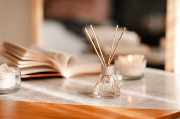 Liquid home fragrance in glass bottle with bamboo sticks staying on marble coffee table over candle and open book in room close up. Aromatherapy.