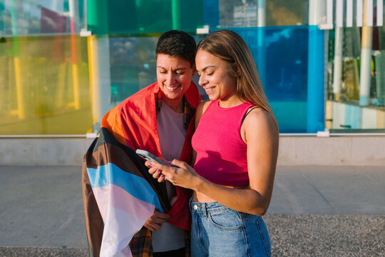 Lesbian Couple Smiling While Looking at a Smartphone with Pride Flag