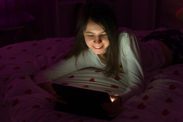 Fototapeta premium Young woman typing on a digital tablet at home. Beautiful girl on the bed in a dark room, lying and holding the electronic device. Smiling happy face, lifestyle and technology concepts.