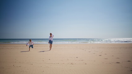Happy boy running mother at sunny beach near ocean waves. Playful child woman
