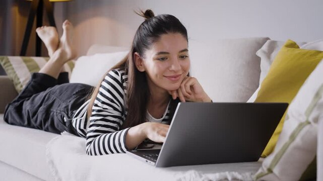 Effort meets comfort as a smiling woman lounges on the sofa, effortlessly working on her laptop. The perfect blend of relaxation, remote work, and modern home vibes