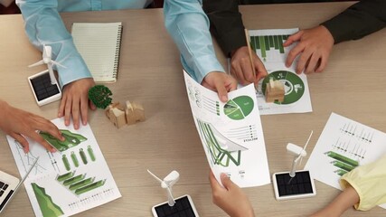 Excellent business team discussing on eco house project at meeting table with windmill design model and green power usage graph for decreasing carbon footprint, save earth. Focus on hand. Delineation.