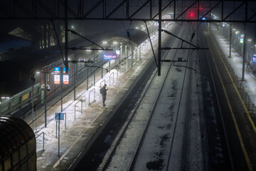 Obraz premium Solitary man on misty platform, shiver in winter air and waiting train. Glow of distant lights, frosted signs and schedule boards, delayed train, cold isolation on dark, solo travel at foggy night