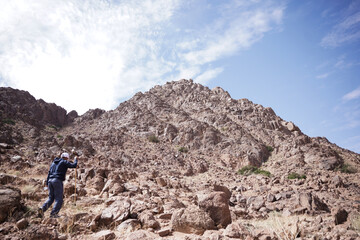A wide view of rugged Saudi desert mountains under a bright sky, with rocky valleys and scattered trees shaping a serene natural landscape. A remote camping spot appears tucked between the cliffs.