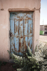 An old Saudi building featuring a weathered blue metal door surrounded by desert plants and sun-faded walls. The rustic textures reflect traditional rural architecture and the charm of abandoned Saudi