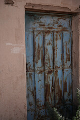 An old Saudi building featuring a weathered blue metal door surrounded by desert plants and sun-faded walls. The rustic textures reflect traditional rural architecture and the charm of abandoned Saudi