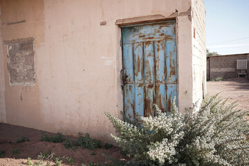 An old Saudi building featuring a weathered blue metal door surrounded by desert plants and sun-faded walls. The rustic textures reflect traditional rural architecture and the charm of abandoned Saudi