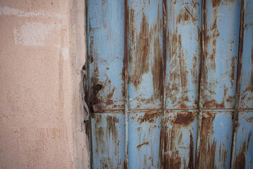 An old Saudi building featuring a weathered blue metal door surrounded by desert plants and sun-faded walls. The rustic textures reflect traditional rural architecture and the charm of abandoned Saudi