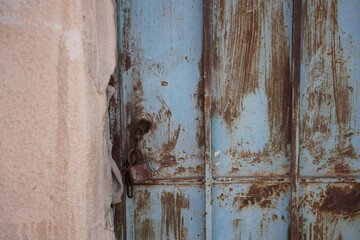Close-up of an old, weathered blue metal door with rust details and a padlock, set against a textured desert wall. A scene reflecting traditional Saudi architecture, age, and authentic rustic charm.