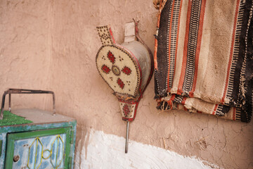 Traditional hand bellows with painted motifs hanging on a mud-brick wall beside woven textiles and a rustic cabinet. A cultural scene reflecting heritage craftsmanship and rural desert d&eacute;cor.