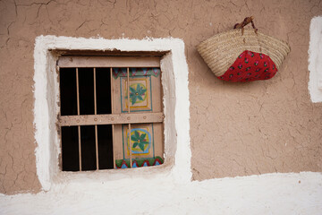 Traditional mud-brick wall with a wooden window featuring colorful painted motifs and a woven basket hanging beside it. A rustic cultural scene highlighting heritage architecture and handmade crafts.