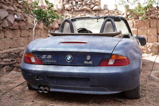 An abandoned BMW Z3 Roadster convertible sits in a dusty yard in Saudi Arabia, surrounded by rustic stone walls. The faded paint and worn-out details highlight the car's age and neglected condition.