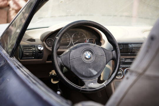 A weathered BMW Z3 Roadster convertible sits abandoned in a rustic yard in Saudi Arabia. The dusty car shows signs of aging, with faded paint and worn-out details, surrounded by old stone walls and na