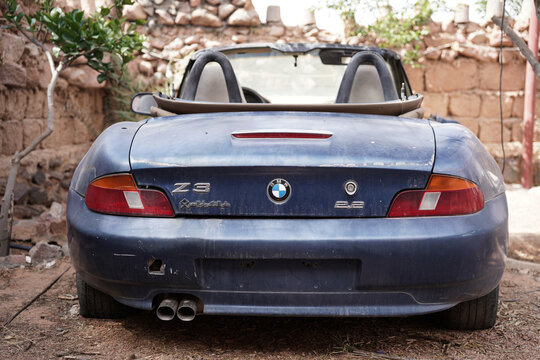 An old and abandoned BMW Z3 Roadster convertible sits in a dusty yard in Saudi Arabia, surrounded by rustic stone walls. The worn-out car shows signs of decay, adding to its nostalgic and vintage char