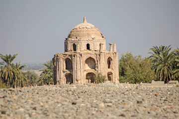 This image depicts a historic place named Bhag Nari in the Balochistan province of Pakistan, a domed structure in a state of ruin, surrounded by lush palm trees and a rocky foreground. 
