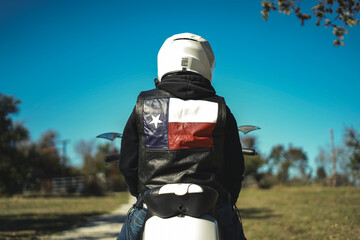 Man Wearing Texas Flag Leather Jacket and Helmet Riding Motorcycle Down Gravel Road With Blue Sky