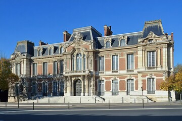 Fototapeta premium A stunning historic mansion, adorned with intricate architectural details, stands majestically under a sunlit blue sky amidst a lively urban setting