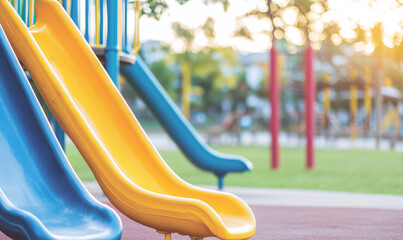 A close-up view of colorful playground slides in a park during sunset. The yellow and blue slides are prominent, with a blurred background of playground equipment and greenery.