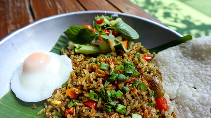 Indonesian fried rice (nasi goreng) with an egg, vegetables and crispy cracker served on a banana leaf in a silver pan