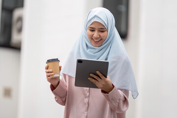 Muslim business woman in hijab holding coffee and using a tablet.