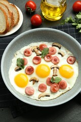 Tasty fried eggs with cut sausages and mushrooms in frying pan served on black table, closeup