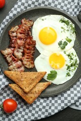 Tasty fried eggs, bacon and pieces of toast on black table, flat lay