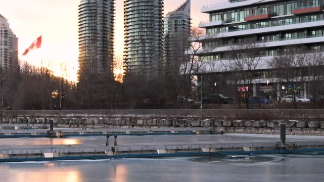 A tranquil sunset at Toronto&rsquo;s waterfront near Mimico and Lake Shore. High-rise buildings glow in the golden light as ducks swim peacefully in the calm water, creating a serene urban scene.

