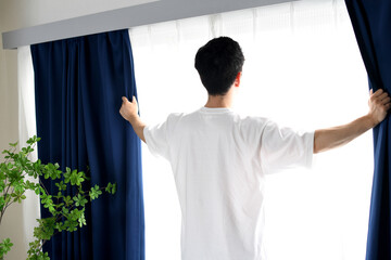 カーテンを開け日光を浴びるアジア人男性