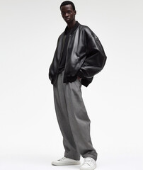 A black male model wearing an oversized leather bomber jacket, baggy trousers and white t-shirt underneath, full body shot, standing straight with legs apart in front of a plain background. 
