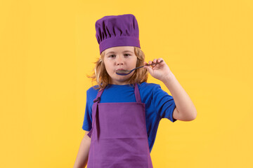 Funny kid chef cook with spoon, studio portrait. Child cooking, little chef prepares food. Kid boy in chefs hat and apron on yellow studio isolated background.