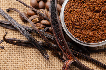 A close-up of roasted coffee beans, dried vanilla pods, and finely ground coffee on a burlap surface. 